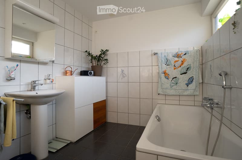 Modern bathroom with white tiles, a sink, bathtub, mirror, and a towel with a fish design. Includes a small plant on the windowsill and a toothbrush holder on the wall.