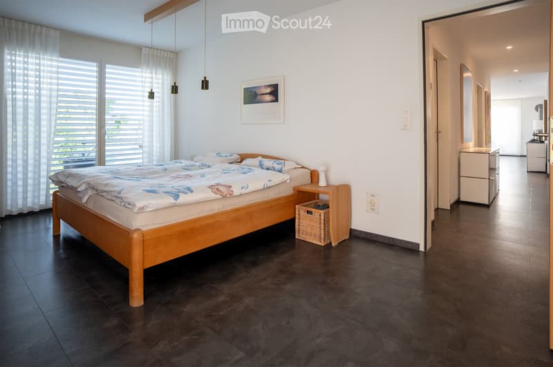 Modern bedroom with a wooden bed, nightstand, wicker basket, and a picture frame on the wall. The room features dark tiled flooring and large windows with white curtains.