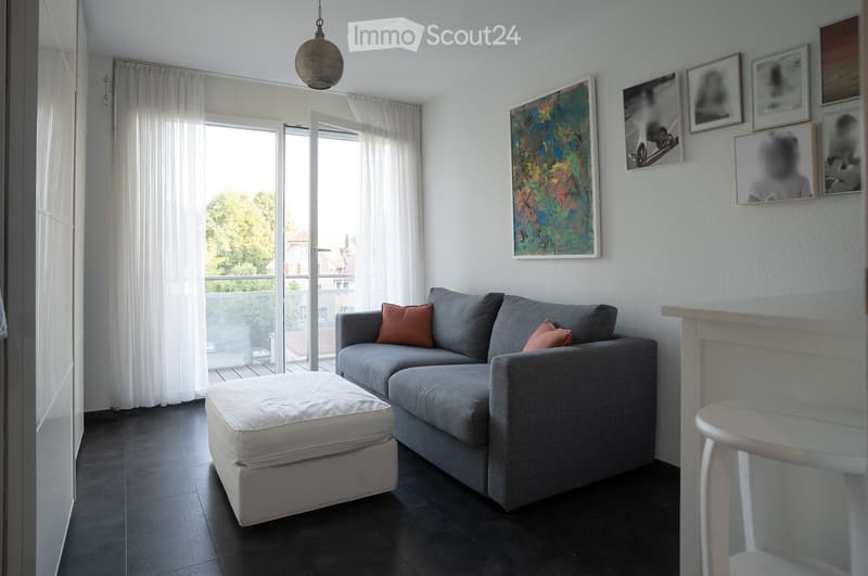 Modern living room with grey sectional sofa, white ottoman, small white stool, colorful painting on wall, large windows with white curtains, and access to a balcony.