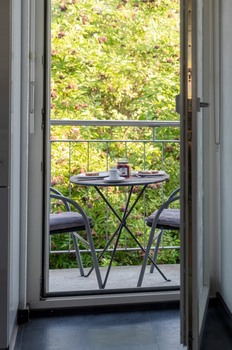Balcony with a small round table, two chairs, a cup, plates, and a jar of jam, overlooking a lush green garden.