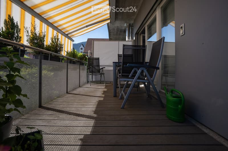 Balcony with wooden decking, metal railing, and a retractable awning. Includes a table, two chairs, and a green garden hose.