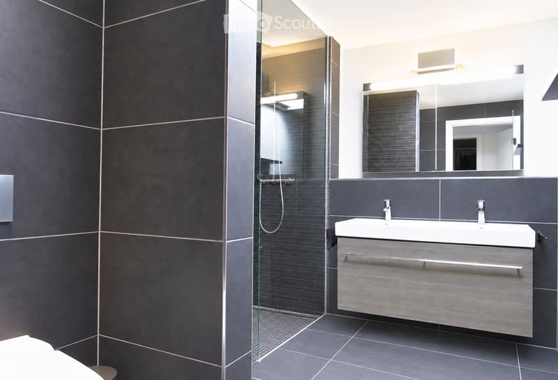 Modern bathroom with dark grey tiles, a large mirror, a double sink, and a glass-enclosed shower area.