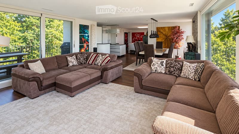Modern living room with large windows offering a view of greenery, featuring a brown sectional sofa, ottoman, dining area with table and chairs, and decorative art pieces.