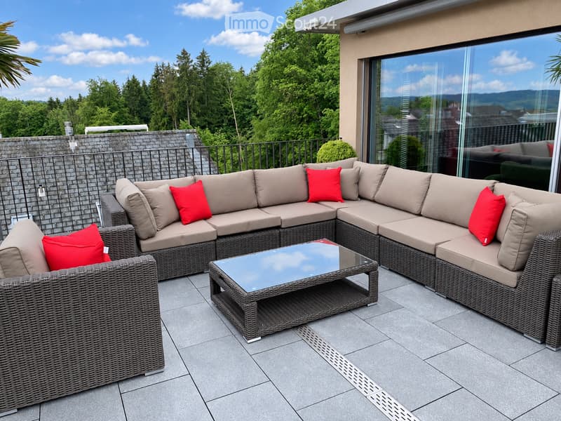 Outdoor patio with a large sectional sofa, coffee table, and red throw pillows, overlooking a lush green landscape.
