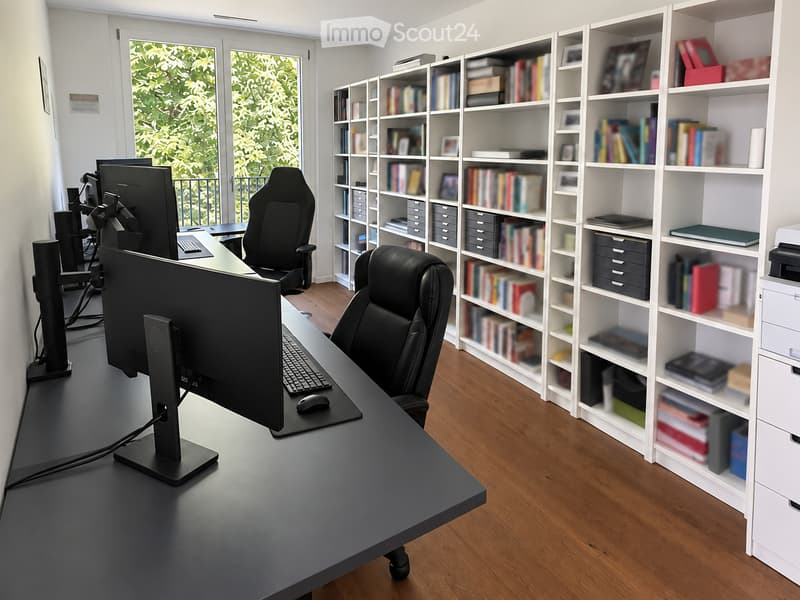 Modern office space with a large desk, ergonomic chairs, dual monitors, keyboard, mouse, and a bookshelf filled with books and office supplies.