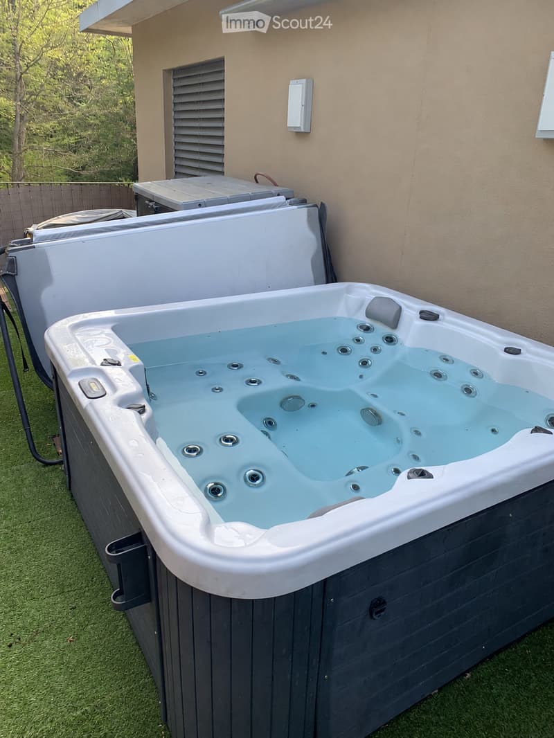A modern hot tub with multiple jets and a control panel, situated on a grassy area next to a house with beige walls and a covered electrical box.