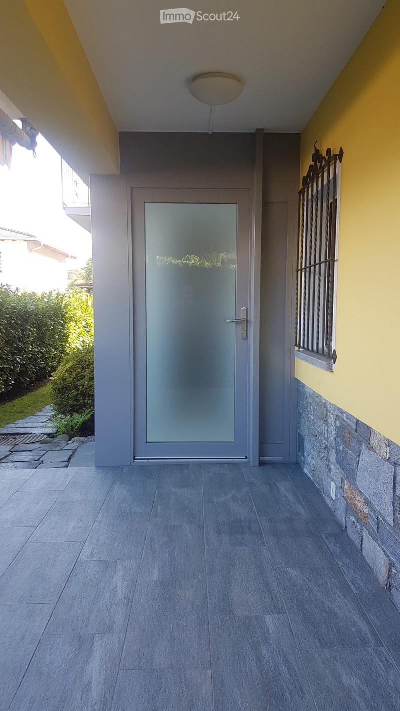 Entrance area with a modern glass door, grey tiled floor, and yellow-painted walls. A small window with decorative metal bars is visible on the right side.