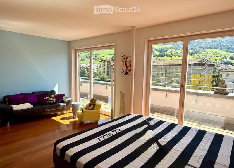 Bright bedroom with wooden flooring, large windows offering scenic views, a bed with black and white striped bedding, a sofa, a yellow armchair, and a small table.