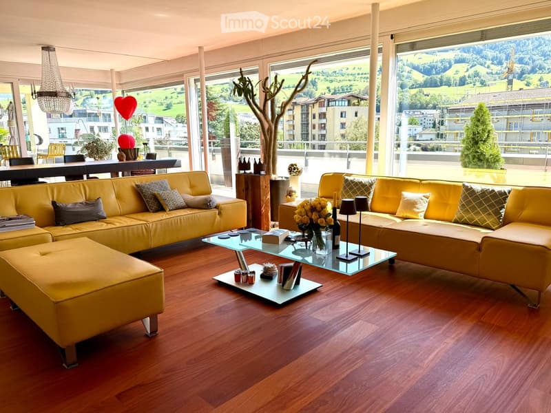 Modern living room with large windows offering a scenic view, featuring yellow leather sofas, an ottoman, a glass coffee table, and a dining area with chairs and a table.