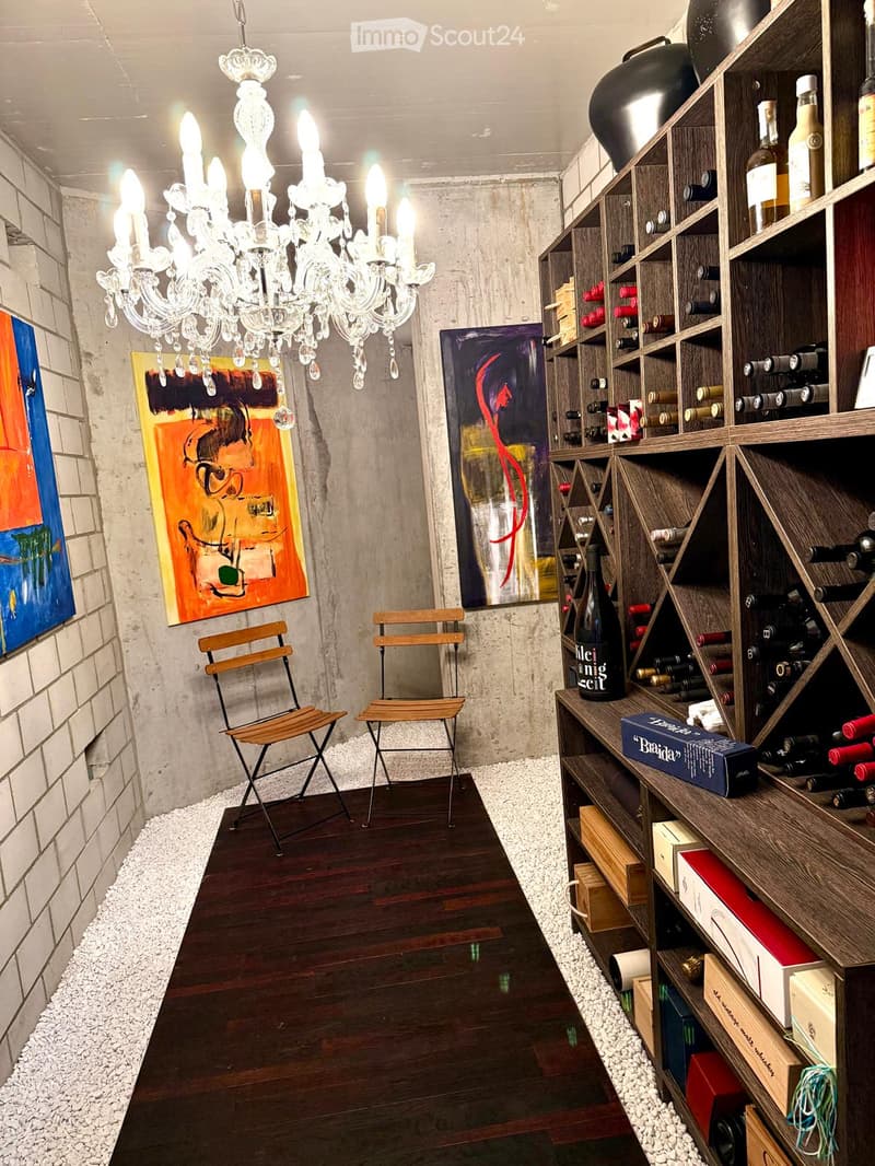 Wine cellar with wooden floor, white gravel accents, chandelier, two wooden chairs, and wine racks filled with bottles.