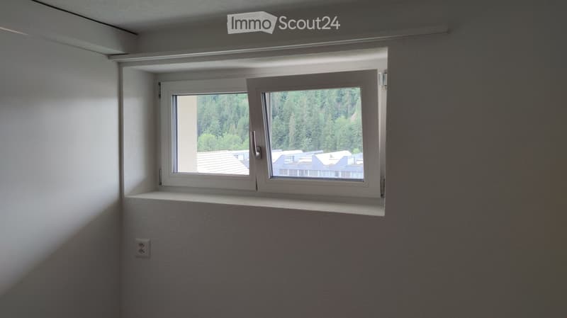 Empty room with white walls and a large window offering a view of trees and rooftops.