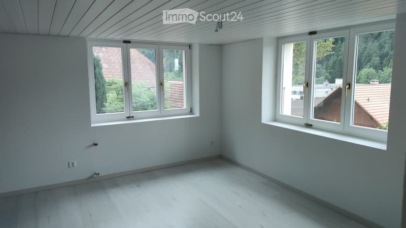 Empty room with light blue walls, white wooden floor, and large windows offering views of neighboring houses and greenery.