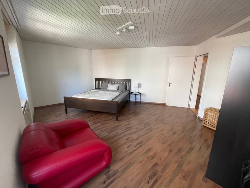 Bright bedroom with wooden flooring, a red leather sofa, a wooden bed with pillows and blankets, a nightstand with a table lamp, and a white door leading to another room.