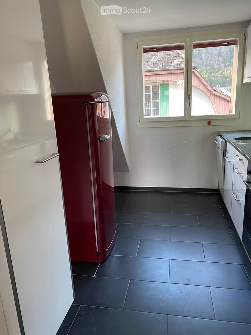 Empty room with dark tiled floor, white walls, and a window with a view of a building and greenery. A red refrigerator is partially visible on the left side.