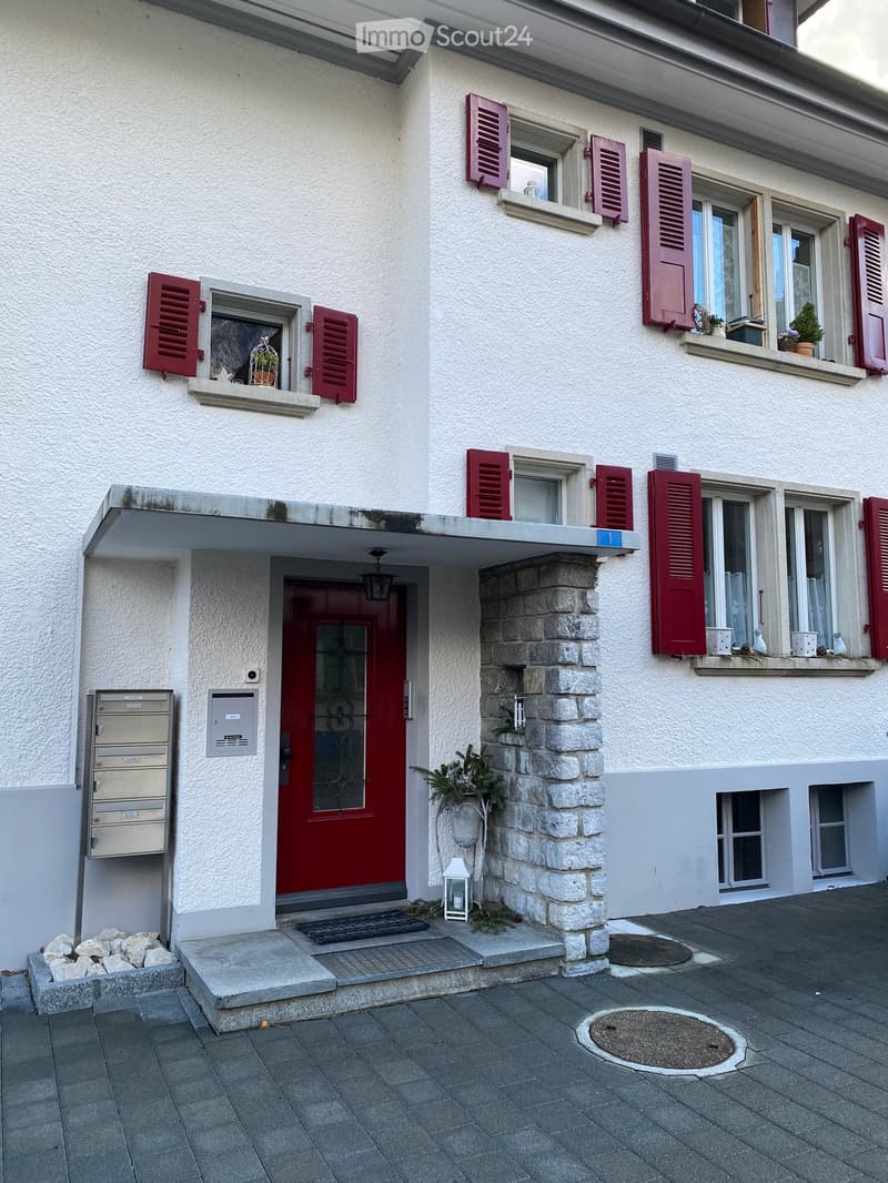 Two-story house with white exterior, red shutters, and a red front door. Features a stone pillar, small garden area, and mailboxes on the left side.