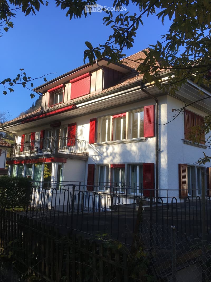 Two-story house with white walls, red shutters, and multiple windows, featuring a fenced front yard and a balcony on the upper level.