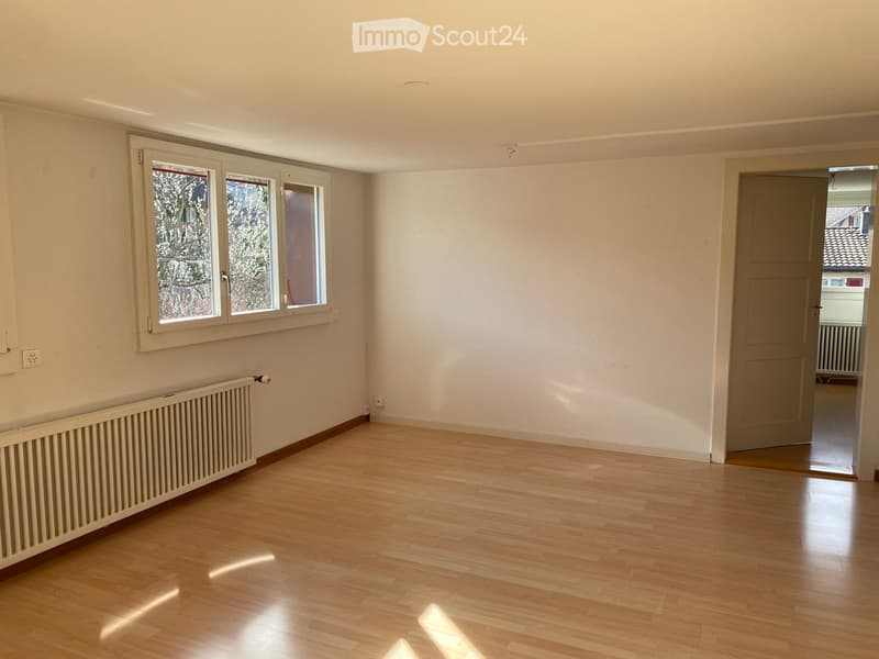 Empty room with wooden flooring, white walls, and large windows with white frames. A radiator is visible along one wall, and a door leads to another room.