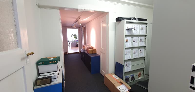 Office corridor with white walls, carpeted floor, and a blue cabinet. Visible items include a printer, a shelf with papers, and several cardboard boxes.