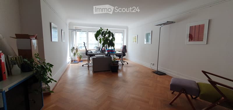 Bright room with wooden flooring, large window, desk with laptop, chair, stool, plant, and floor lamp.
