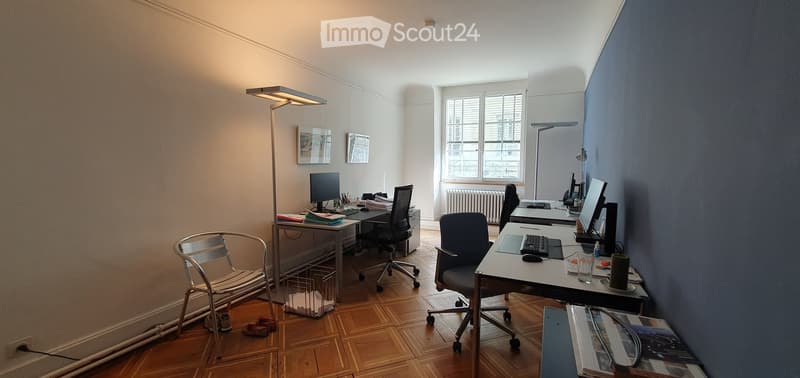 Bright office space with wooden flooring, two desks, chairs, computers, and lamps. The room features large windows and a mix of modern and classic decor.