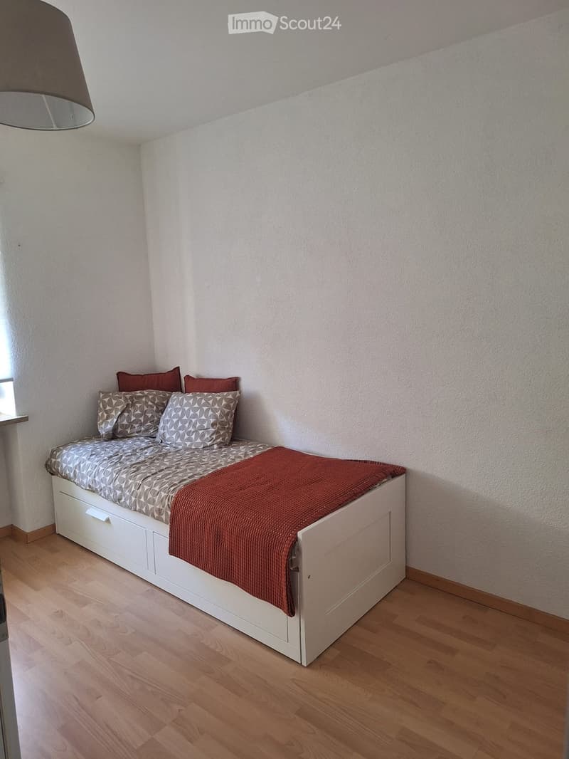A small, cozy bedroom with a white bed featuring a geometric-patterned duvet and red throw, placed against a white wall with wooden flooring.