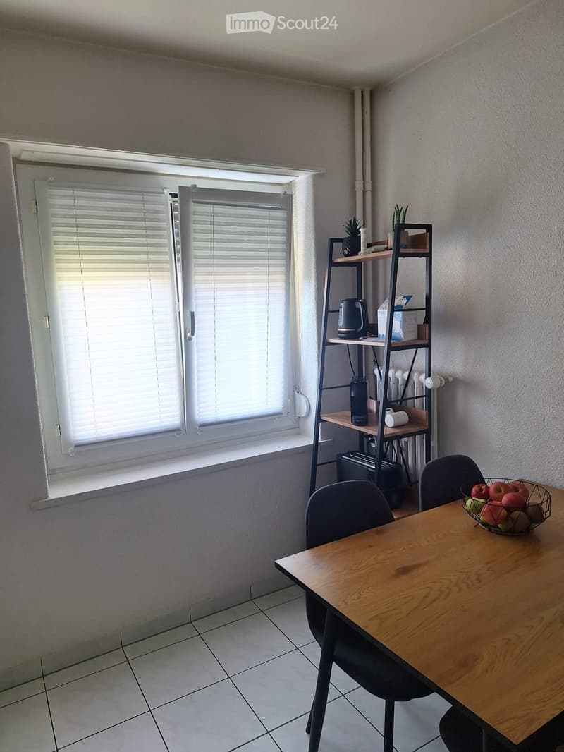 A small, well-lit room with a wooden dining table, two chairs, a shelf with a kettle and other items, and a fruit bowl on the table. The room has tiled flooring and a window with blinds.