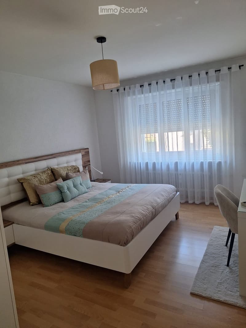 Modern bedroom with a large bed, multiple pillows, a nightstand, a chair, a small table, and a white rug on the wooden floor.