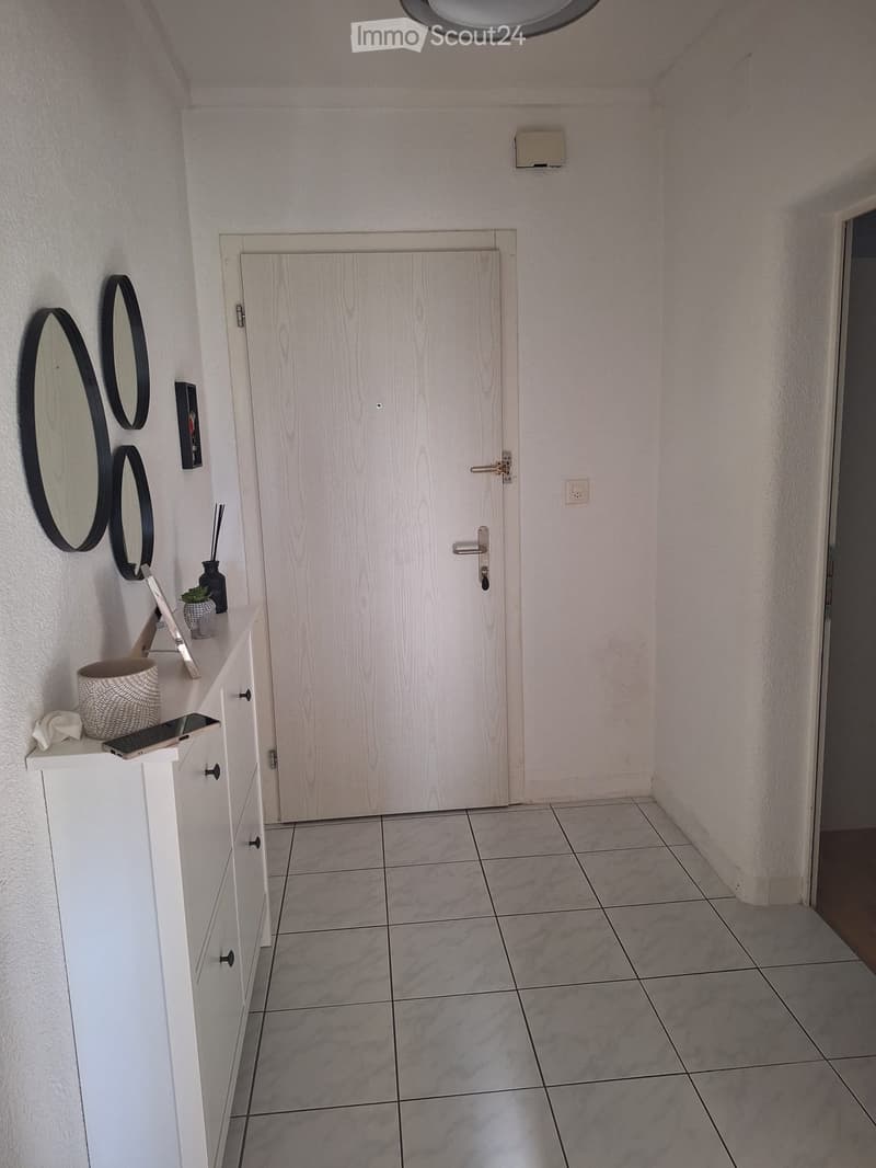 A clean, modern hallway with tiled flooring, white walls, and a white wooden door. A small white cabinet with drawers is positioned on the left, holding decorative items including a woven basket, a small potted plant, and a framed picture. Three round bla