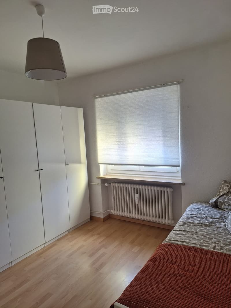 A small, well-lit bedroom with wooden flooring, a single bed with patterned bedding, a white wardrobe, a radiator, and a window with a blind.