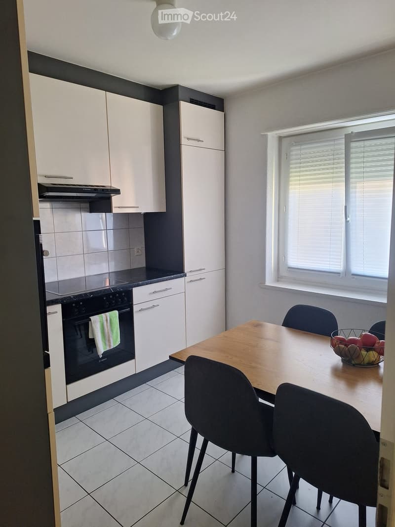Modern kitchen with white cabinets, black countertops, a built-in oven, a dining table with chairs, and a fruit bowl on the table.