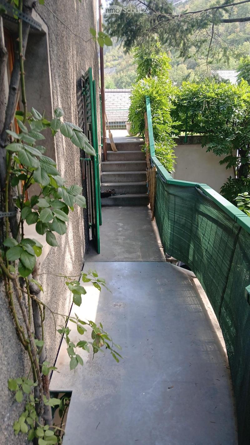 A narrow concrete pathway bordered by green railings, leading to a set of stairs and a small outdoor area with plants and a view of greenery.
