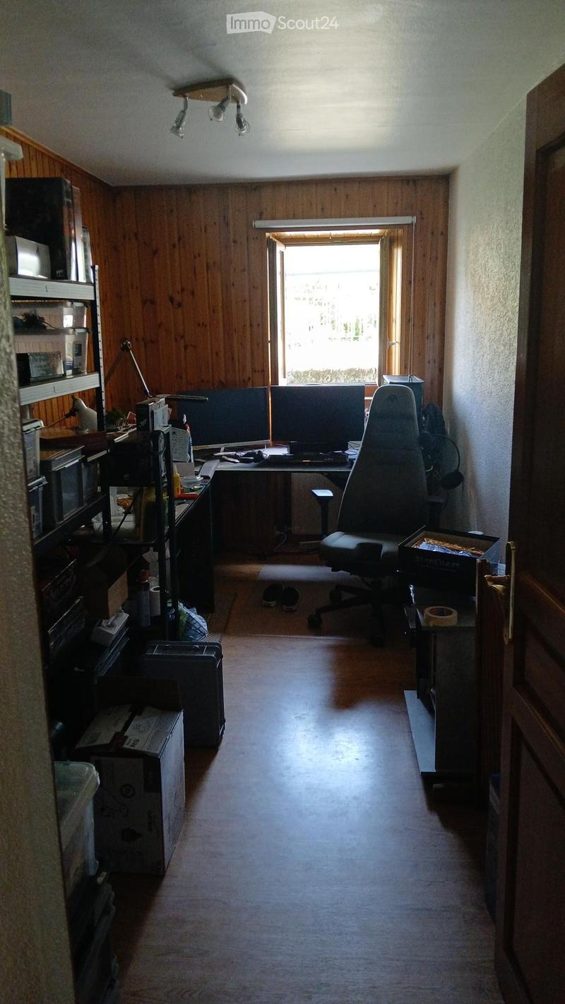 A furnished room with wooden walls, a large window, a desk with two screens, a chair, shelves, and various items scattered around.