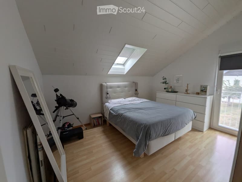 Bright bedroom with wooden flooring, a large bed with grey bedding, a nightstand with a lamp, a telescope on a stand, and a large mirror leaning against the wall.