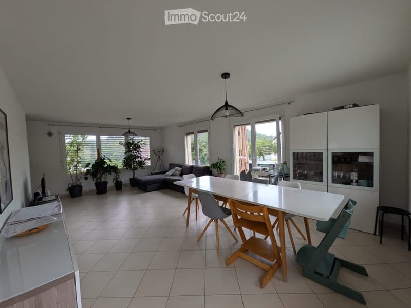 Bright living and dining area with a grey sectional sofa, white dining table, and modern chairs. Large windows and potted plants add to the space.