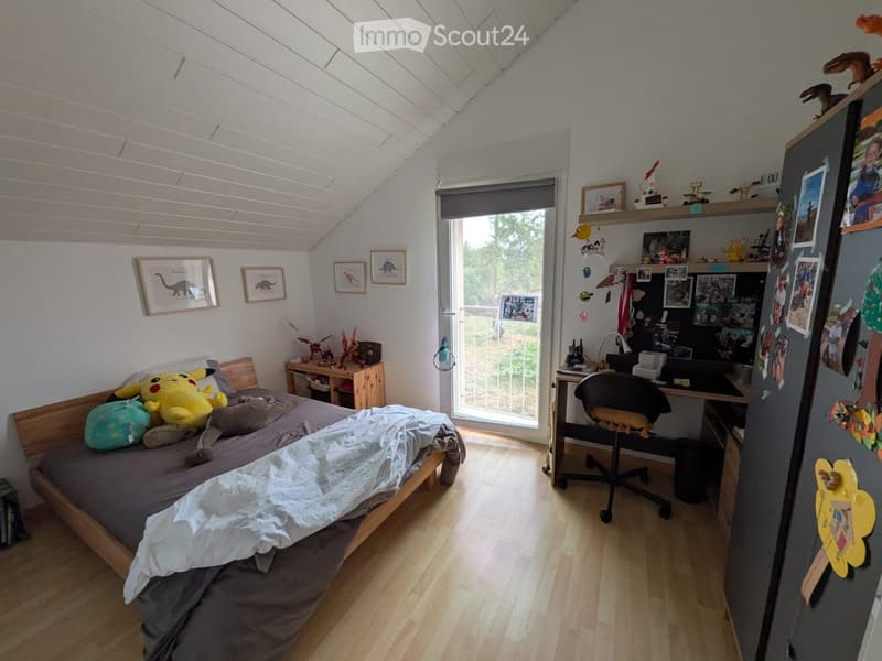 Cozy bedroom with a slanted ceiling, wooden floor, and large window. Features a bed with pillows and blankets, a desk with a chair, a fridge with photos and magnets, and shelves with toys and framed pictures.