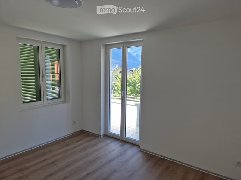 Empty room with wooden flooring, white walls, and large windows, including a sliding glass door leading to a balcony.