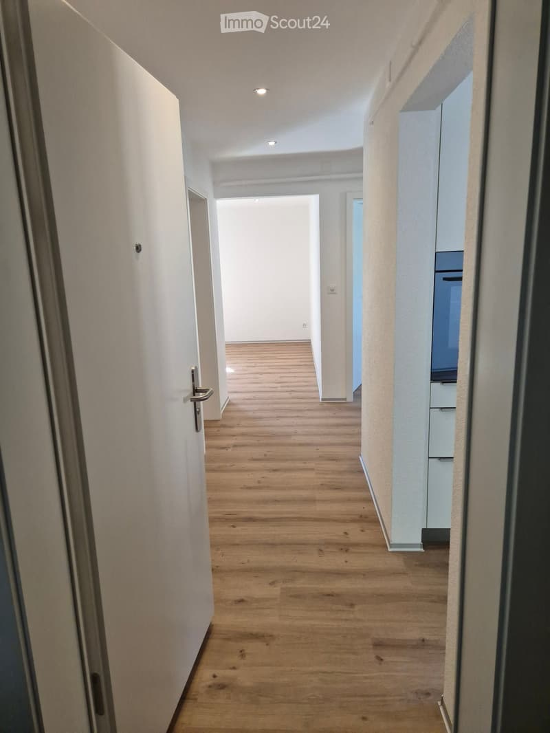 A clean, modern hallway with light wooden flooring, white walls, and recessed ceiling lighting. The hallway features a door on the left and an open doorway leading to another room on the right.