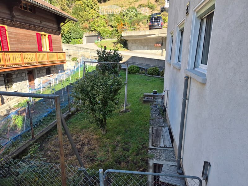 Garden area with grass, small tree, fence, and steps leading to a house entrance. A raised ground floor is visible, with a wooden deck and a small utility area near the house.
