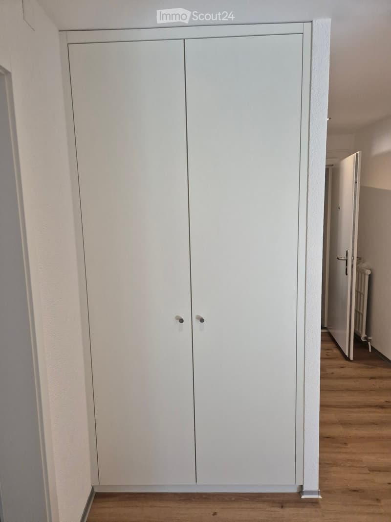 Empty hallway with white built-in wardrobes on one side and wooden flooring, leading to another room with a radiator visible in the background.