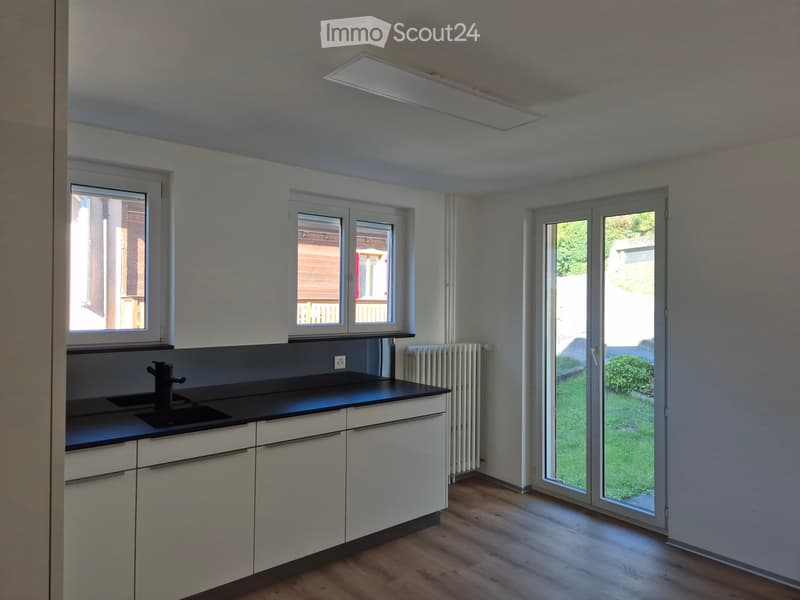 Modern kitchen with white cabinets, black countertop, wooden floor, and large windows. Features a sink with a black faucet and a radiator under one window.