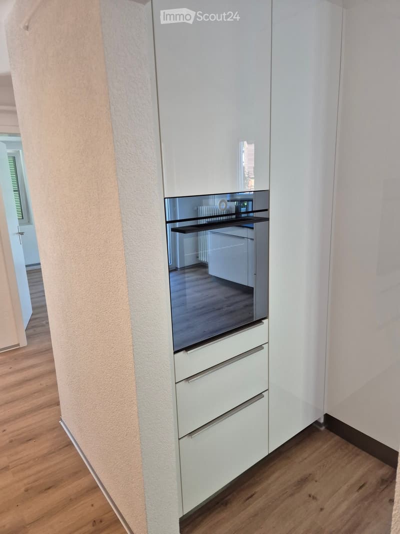 Modern kitchen with white cabinets, a stainless steel refrigerator, and an oven, featuring wooden flooring and a visible hallway.