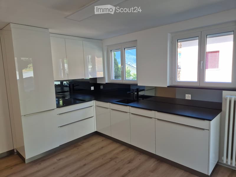 Modern kitchen with white cabinets, black countertop, and wooden flooring. Features a sink with a faucet and large windows offering a view of greenery.