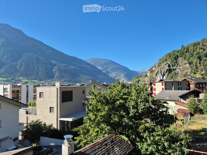 A scenic view of a town nestled in a mountainous valley, with modern and traditional buildings, lush greenery, and a clear blue sky.