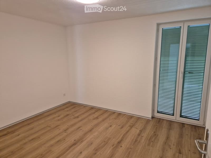 Empty room with light-colored walls and wooden flooring, featuring a large sliding glass door with blinds.
