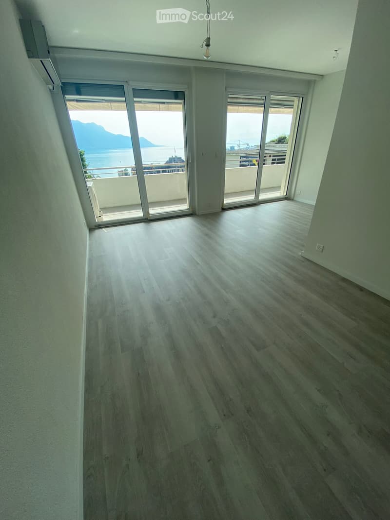 Empty room with wooden flooring, large sliding glass doors leading to a balcony with a scenic view of a lake and mountains.