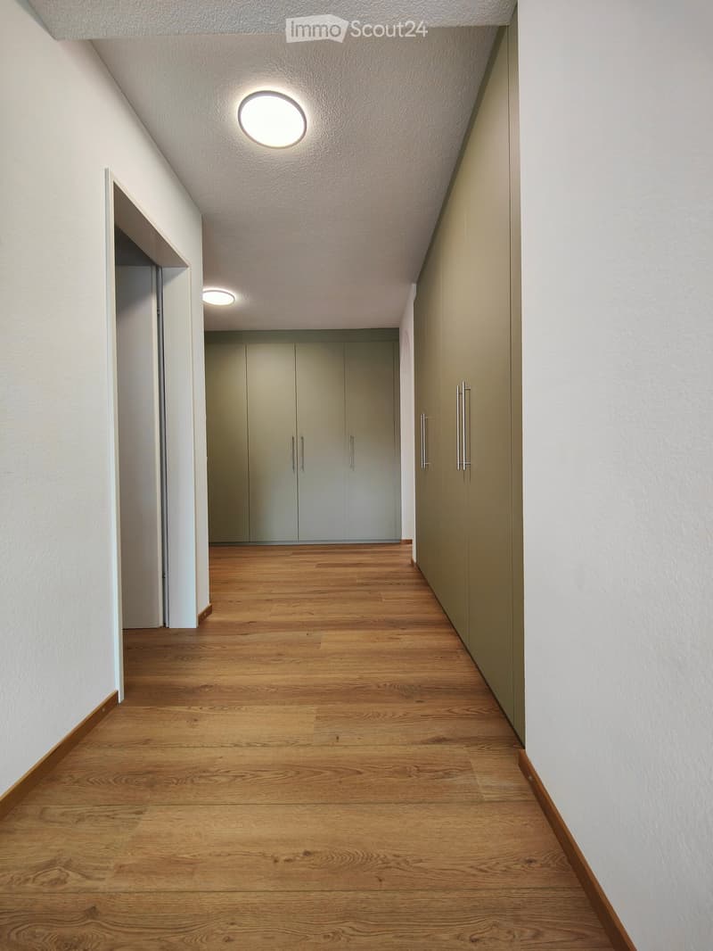 Modern hallway with wooden flooring, white walls, and recessed lighting. Features built-in storage units with sleek metal handles on one side and an open doorway leading to another room.