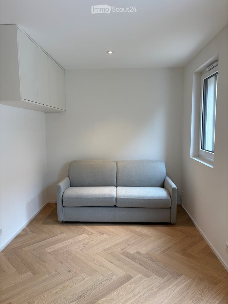 A modern, minimalist living room with a grey sofa, light wooden herringbone flooring, white walls, and a built-in white cabinet. A small window is visible on the right side.