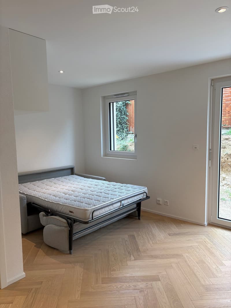 Minimalist bedroom with a grey sofa, a bed, wooden flooring, and large windows offering a view of greenery.