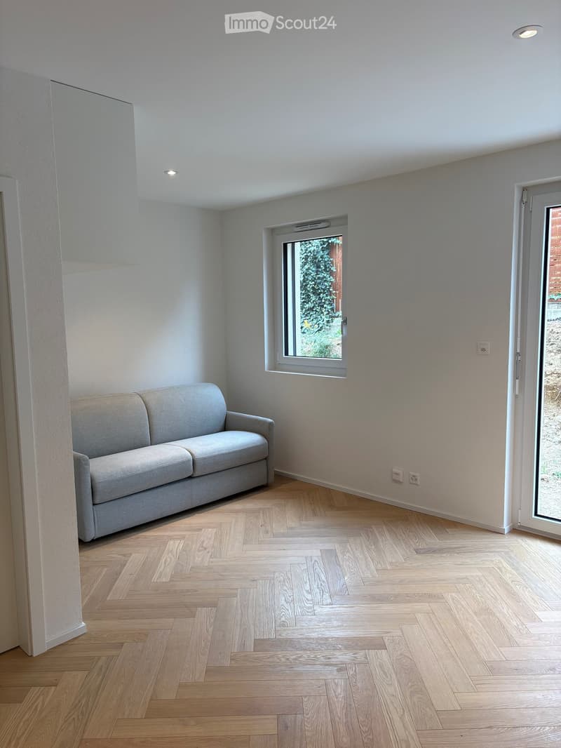 Living room with light grey sofa, wooden herringbone floor, large window, and sliding glass door leading outside.
