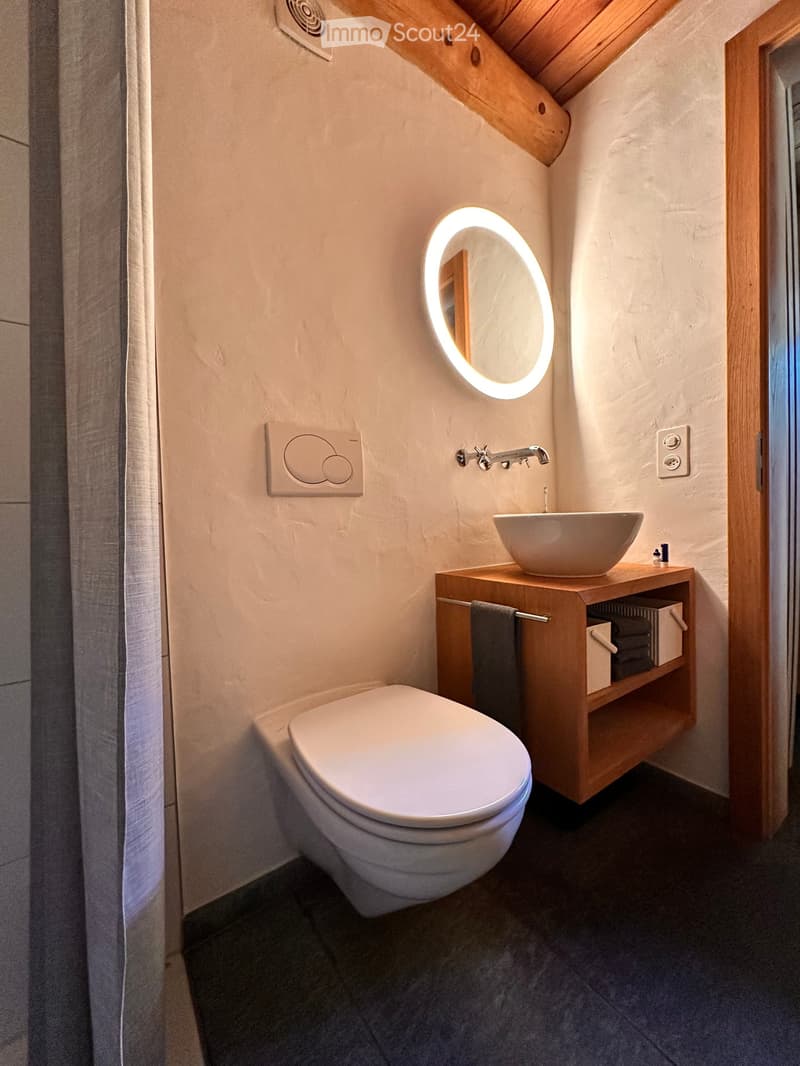 Modern bathroom with a toilet, wooden sink cabinet, round illuminated mirror, and towel on a shelf.